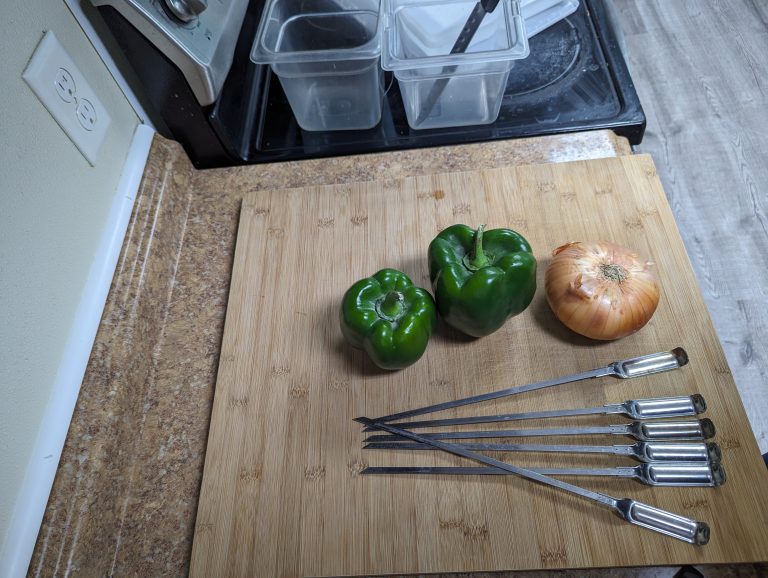 A wooden cutting board on a kitchen countertop with two green bell peppers, a yellow onion, and six stainless steel skewers arranged neatly.