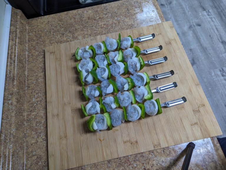 A set of assembled shrimp kebobs on a wooden cutting board, each skewer alternating raw shrimp, green bell peppers, and onions.