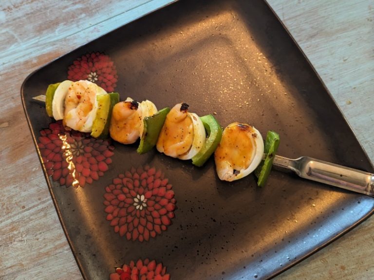 A grilled shrimp kebob with vibrant green peppers, tender onions, and shrimp marinated in Peruvian Gold sauce, served on a decorative square plate with floral designs.